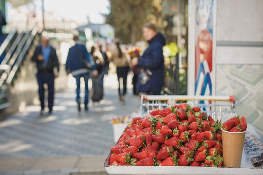 Fresh ripe red strawberries sold in paper cup at street market stall with blurred pedestrian background on sunny day