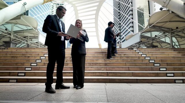International business professionals from diverse ethnic backgrounds standing together on city steps, discussing ideas and using digital devices in an urban setting