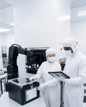 Engineers in cleanroom suits monitoring robotic arm handling silicon wafer with tablet