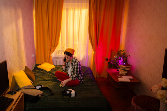 Young girl in casual fashion sitting on her bed and enjoying phone talk. Cozy home interior and connection in relaxing atmosphere. Wide shot