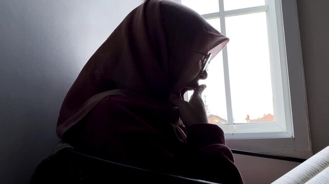 Slow motion low angle side view of an Asian Muslim woman in a hijab and glasses. She is sitting indoors reading a book by a bright window, enjoying a quiet and peaceful study time at home.