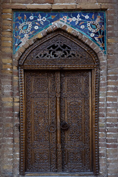Carved wooden door of Gur-e-Amir mausoleum in Samarkand with intricate floral patterns and blue mosaic tilework above. Ideal for travel publications, heritage content, and cultural design projects.