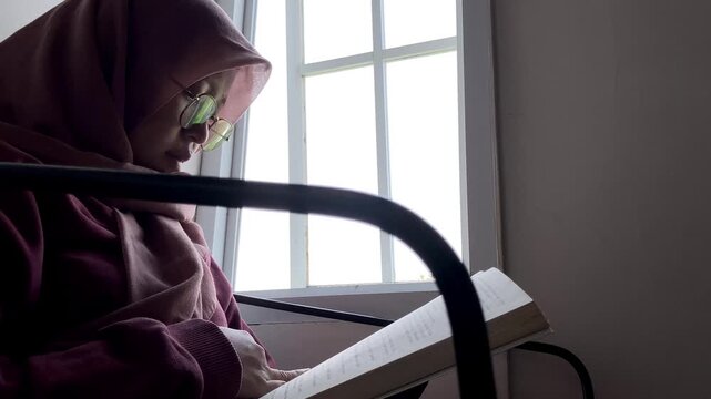 Slow motion low angle side view of an Asian Muslim woman in a hijab and glasses. She is sitting indoors reading a book by a bright window, enjoying a quiet and peaceful study time at home.