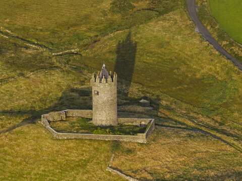 Aerial view of Doonagore Castle, a historic stone tower house with a low stone wall in a grassy field and a long shadow cast by the sun in Doolin, County Clare, Ireland.