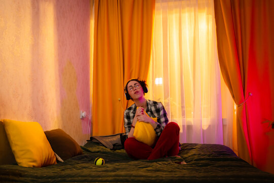 Girl listening to pop music and moving joyfully on her bed in sunset-lit room. Teen leisure, home comfort and emotional wellness. Wide shot, low angle view