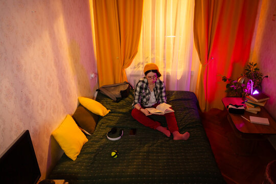 Woman in relaxed clothing focuses on reading while seated on bed in golden hour light. Calm mood, cozy interior, and personal development through books. Wide shot