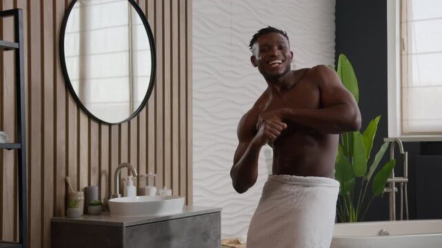 Happy African American man dancing in bathroom with bath towel on hips listening to music pointing finger at camera smiling joyful playful hipster male guy having fun morning hygiene routine indoors