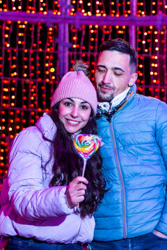 Young couple enjoying christmas lights and holiday season, holding heart lollipop