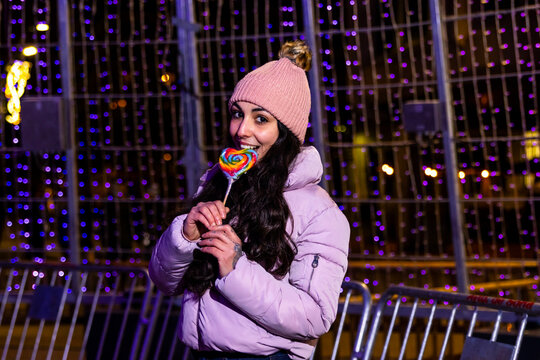 Young woman wearing a winter jacket and hat, holding a rainbow lollipop at night