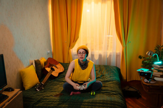 Caucasian teenager in a striped shirt and beanie is gaming with a controller at home during sunset. Concept of digital play and adolescent relaxation. Wide shot