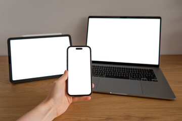 Hand holding smartphone with blank screen in front of laptop and tablet, representing digital technology, mobile communication and connected devices © Anna