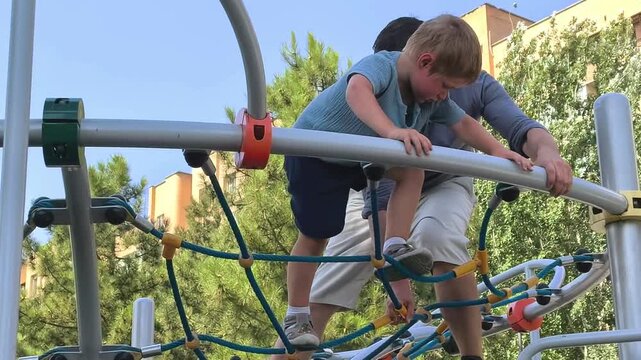 Father helps his young son climb playground equipment outdoors, active childhood, parental support, family bonding and summer recreation.