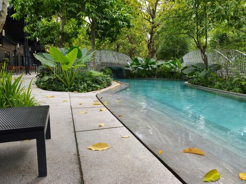 A low-angle shot of a serene outdoor swimming pool designed to resemble a natural river channel, surrounded by lush green tropical plants, foliage, and small decorative footbridges in a resort setting
