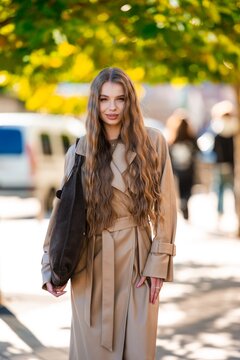 woman in stylish trench coat standing outdoors on urban street with trees and vehicles, casual and fashionable, beige and brown tones, fabric and leather materials, front view.