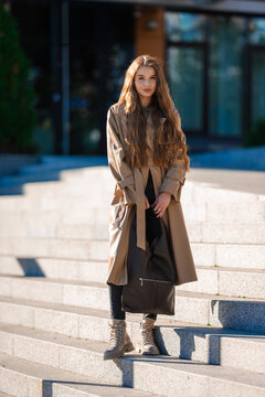 young woman standing confidently on urban steps in trench coat and black bag, front view, fashion street style, beige and tan tones, confident and stylish mood.