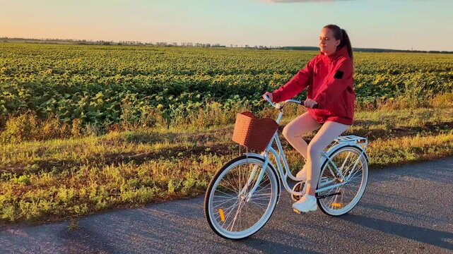 A woman cycles on a road next to a large sunflower field with the sun setting in the background. The scene shows a calm evening in a rural setting