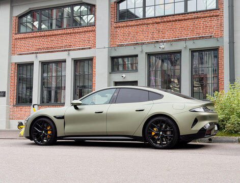 Matte olive green Xiaomi SU7 electric sports sedan car wrapped in protective PPF film parked near loft red brick building facade, contemporary Chinese EV luxury automobile paint protection wrap.