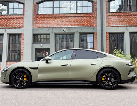 Matte olive green Xiaomi SU7 electric sports sedan car wrapped in protective PPF film parked near loft red brick building facade, contemporary Chinese EV luxury automobile paint protection wrap.