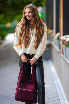 woman smiling and holding a bag in an outdoor urban setting with flowers in the background, happy and stylish front view, beige and black leather pants, suede bag, fabric jacket, fashion.