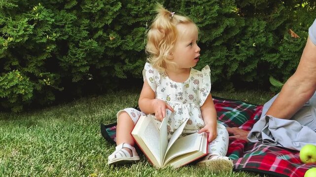 Toddler girl turns pages of a book during an outdoor picnic, early learning, childhood reading, family leisure and summer education.