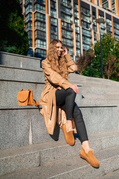 woman casually sitting on urban steps with tan coat and black leggings, side view, modern building background, fashion and lifestyle context, casual and relaxed mood.