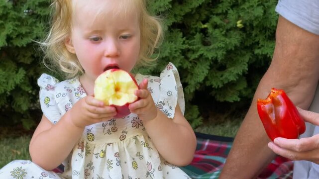 Young child holds and eats a fresh red apple outdoors, healthy snack, childhood nutrition, fresh fruit and natural summer lifestyle.