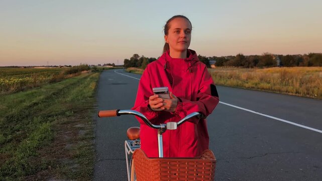A woman stands next to her bicycle on a quiet road. She is looking at her smartphone while the sun sets in the background, creating warm colors in the sky