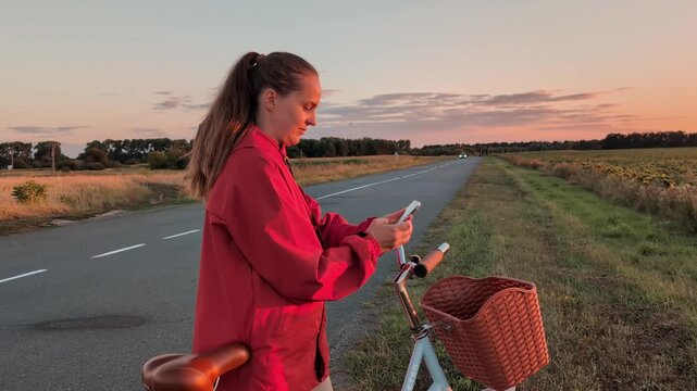 A woman in a red jacket stands by her bicycle on a country road at sunset. She looks at her phone while fields stretch out on either side