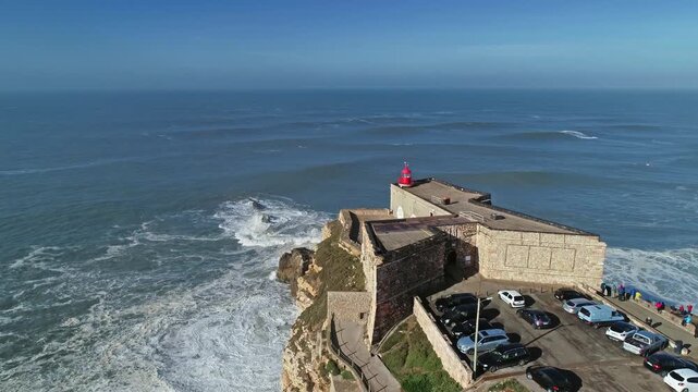 Aerial view of the historic Forte de Sao Miguel Arcanjo lighthouse on rocky cliffs overlooking the Atlantic Ocean with parked cars in Nazare, Leiria District, Portugal.