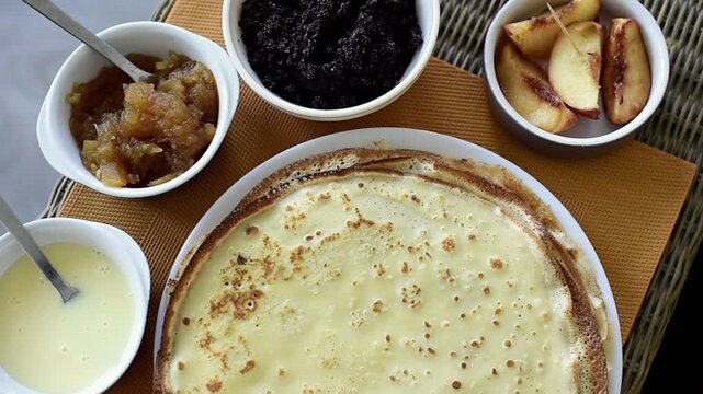 A plate of thin, golden-brown pancakes sits surrounded by small bowls containing poppy seed filling, fruit jam, condensed milk, and sliced fruit. The breakfast table arrangement rests on a textured pl