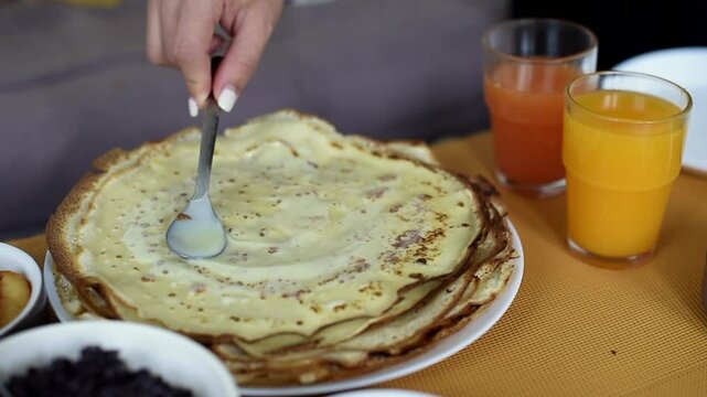 A person holds a spoon to spread sweetened condensed milk across the surface of a thin pancake. Two glasses of fresh orange and grapefruit juice stand on the table nearby.