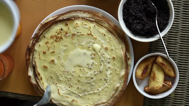 A person uses a spoon to spread sweetened condensed milk across the surface of a golden pancake. Small bowls of poppy seed paste and sliced fruit sit on the table alongside the plate.