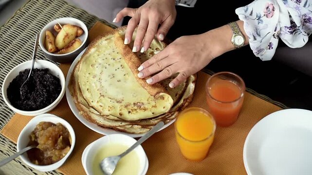 A person uses both hands to roll up a thin, cooked pancake on a plate. The breakfast table is set with small bowls of jam, poppy seeds, condensed milk, fruit, and citrus juice.