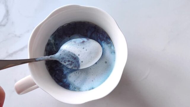 Woman's hand lifts a teaspoon with milk foam from blue matcha in a white cup, healthy morning drink in sunlight.