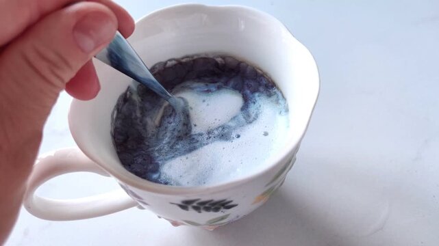 Woman's hand stirs blue matcha in a white cup with a teaspoon on a white table, shown from a side angle, healthy morning drink.