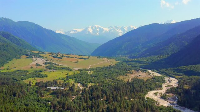 Dolra Valley stretches through aerial view as the drone advances above meadows, forest and a winding river near Mazeri, Svaneti, supporting ecotourism, trekking routes and Georgian travel promotion.