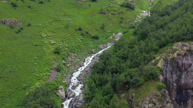 Alpine stream winds in ascending aerial view across green slopes below Shdugra Waterfall near Mazeri, Svaneti, supporting trekking tourism, watershed research, mountain geography and travel media.