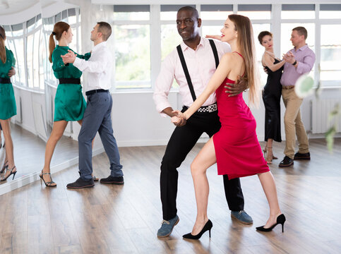 Woman with African man participants of dance workshop perform figure waltz. Students learn to dance classic salon dance, work out movements with partner in choreography studio.
