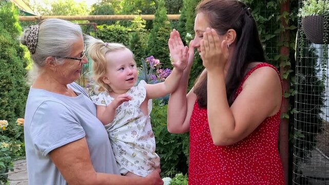 Mother plays peekaboo with a toddler girl while grandmother holds her outdoors, three generation family bonding, joyful childhood and playful affection.