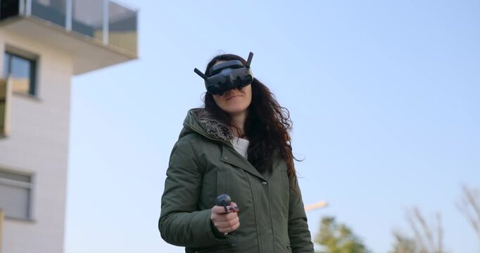 Young woman in a green jacket flies an FPV quadcopter using goggles and a single-hand controller under a clear blue sky.