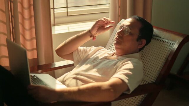 Senior Indian male experiencing eye fatigue after prolonged computer use while sitting near a sunlit window at home.