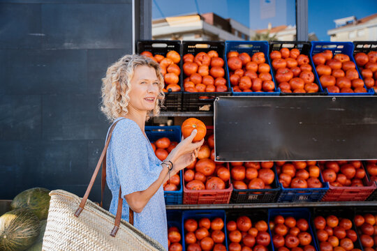 Smiling Mature Blonde Woman Holding Fresh Tomato at Local Produce Market in Spain