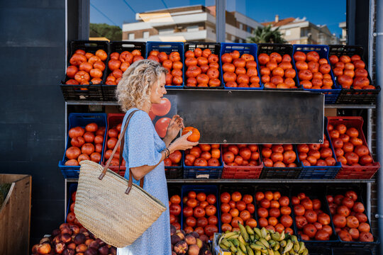 Mature Blonde Woman Choosing Local Tomatoes at Outdoor Spanish Market with Woven Shopping Basket