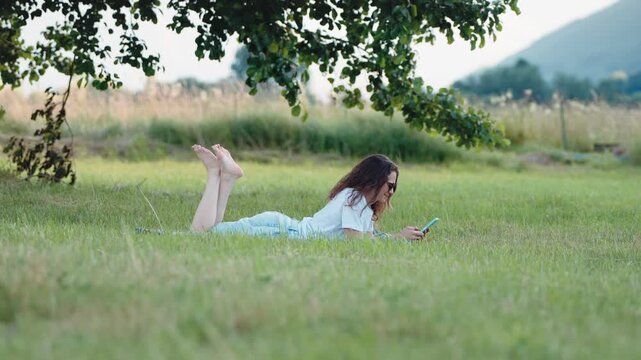 Young woman lying on her stomach on the grass and using a smartphone under a large tree, enjoying a relaxed outdoor moment in nature.