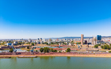Naklejka premium View of a riverside city with buildings and cranes under a clear blue sky. Cities line the river with boats and cranes. Clear blue sky above shows a bright day.