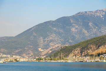 Fototapeta premium Scenic Fethiye town and marina under warm evening light and mountain haze in Turkey. Mediterranean coastal resort landscape, yacht masts, calm sea water, and sunset atmosphere.