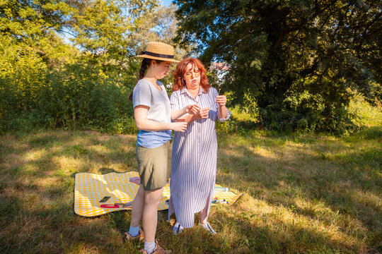 Mother and adult daughter enjoy a summer park picnic sharing laughter and fun while surrounded by vibrant grass and trees in a calm outdoor environment.