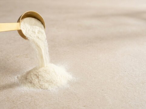 Collagen protein powder pouring from golden measuring scoop onto neutral surface