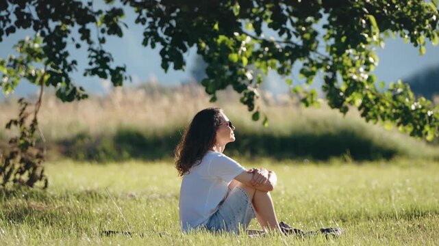 Young woman relaxing while sitting on the grass under a large tree, enjoying a peaceful outdoor moment, fresh air and connection with nature.