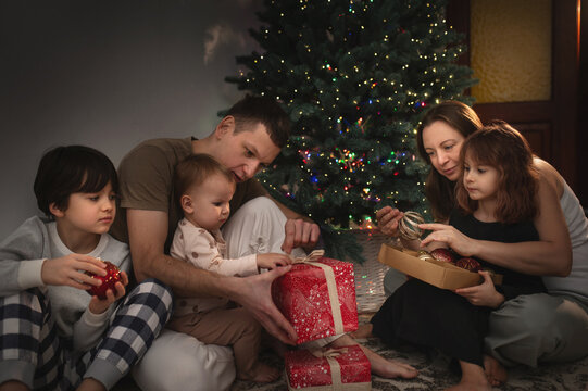 A large family of three children, mom and dad, are celebrating New Year's and Christmas under the Christmas tree at home, opening presents. A quiet, cozy family evening.